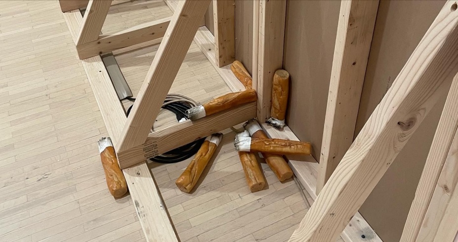 Eight oversized cigarette butts are huddled behind a set-like temporary wall made of unpainted, light-colored 2x4 wood framing studs and unpainted sheetrock. The color of the studs matches that of the wooden floor. Golden baguettes stand in for the cigarette filters; painted white, their tips are burned. Beige and brown electronic round cables are neatly coiled and tucked under a stud. The image is a close-up that does not reveal the size of the wall or its position in space. Entitled Mégot—or Cigarette Butt, in English--this grouping of sculptures was made by Jean-Charles de Quillacq. The unfinished wall is part of Bridget Moser’s installation When I Am Through With You There Won’t Be Anything Left. 