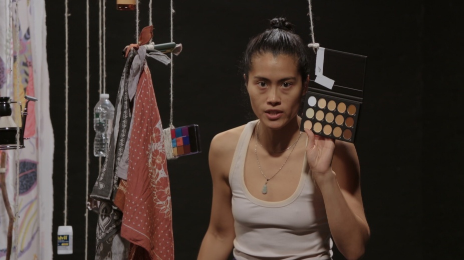 A person wearing a white tank top stands amongst household, cosmetic and pharmaceutical items dangling from strings. This is a still from Every Ocean Hughes’s video entitled One Big Bag. 