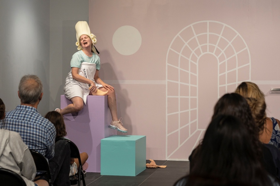 A white, blond-haired woman straddles a waist-high lilac pedestal in front of a rose-gold background that matches the color of her sneakers. An abstract architectural outline and a circle, made to represent the sun, are painted in white on this background. Her head is lightly tilted back, her eyes are closed and her mouth slightly opened. Her facial expression combines smugness, enjoyment, and jouissance. She holds a white skin-toned rubber foot in her right hand. It rests on the pedestal, between her legs. She wears a funny wig, made of beige foam, which recalls the wigs worn by British barristers or European aristocracy. A white towel with undecipherable black text is wrapped over a t-shirt whose mint color matches the small square pedestal below her left dangling foot. She is in front of a white audience of different genders and ages, seen from the back. This is an image from Bridget Moser’s live performance entitled “When I Am Through With You There Won’t Be Anything Left.”. 