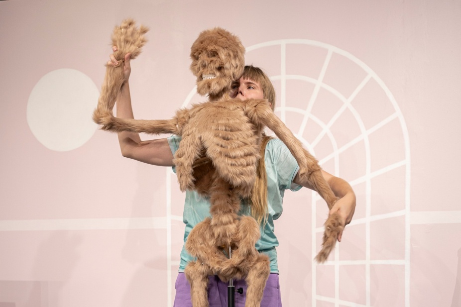 A blond-haired white woman stands behind a furry beige-rose skeleton, holding up its hands. A segment of black pole can be seen at the bottom of the image, below the skeleton’s pelvis: it is part of a structure that holds up the skeleton. The woman wears a mint-colored t-shirt and lilac pants. Her hair is styled with a fringe. She stands in front of a pink wall on which a white abstract architectural outline and a sun have been painted. This is an image from a performance by Bridget Moser entitled When I Am Through With You There Won’t Be Anything Left. 