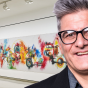A headshot of Anthony Billoni, a white man wearing black rimmed glasses with gray short hair. He is pictured in front of an artwork by FUTURA 2000 in the UB Anderson Gallery. 