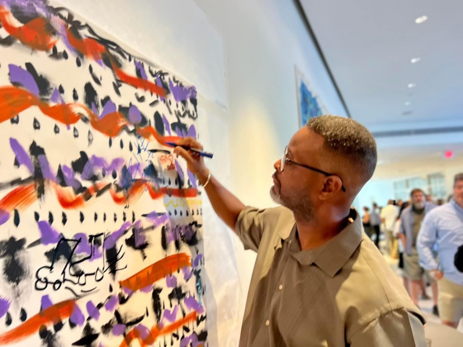 In the foreground a Black man holds a pen to a painting on a wall, while people watch in the background. 