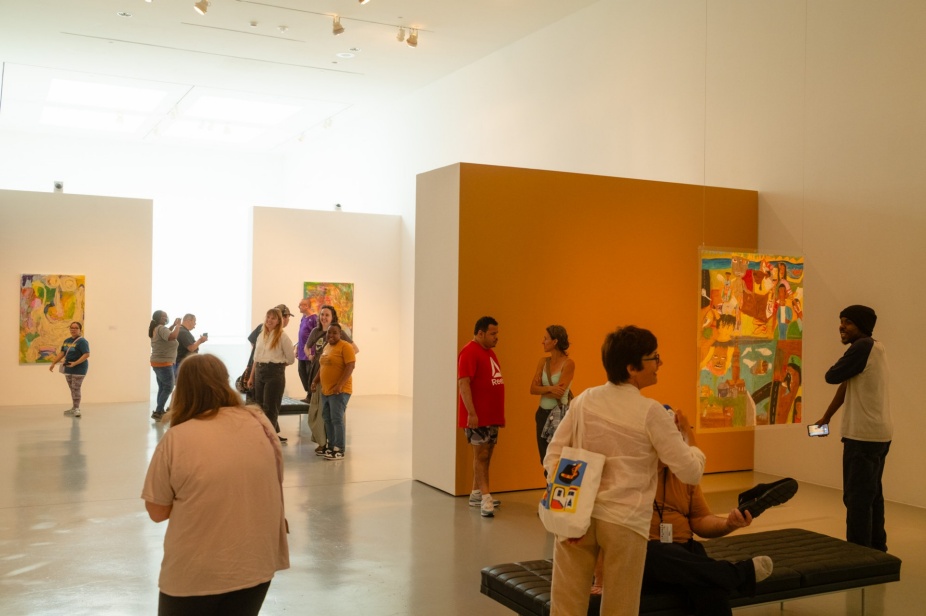 Visitors gathered during the opening of Bits by Bits: Six Artists from Buffalo’s Progressive Studio at the University at Buffalo Art Galleries. People are standing and talking in the gallery, looking at colorful paintings on the walls and a large work displayed on a freestanding orange panel. 