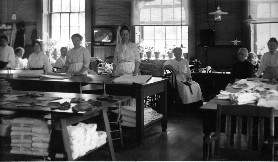 Black and white photograph of a group of women in a laundry room at the Buffalo State Asylum. The women, dressed in light-colored uniforms and dresses, stand or sit at worktables covered with folded linens and fabric. Sunlight streams in through large paned windows at the back of the room, where rows of potted plants line the windowsills. The space is orderly and filled with wooden tables, chairs, and sewing equipment. The overall mood is quiet and industrious. 
