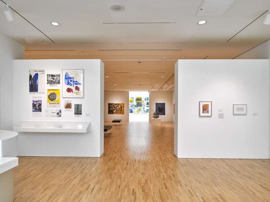 Installation view of Martha Jackson in Paris in the first-floor gallery at the University at Buffalo Art Galleries, CFA Gallery. The photo shows white walls and wood floors with posters and prints on the left wall and framed works on paper on the right. Through the central opening, additional paintings and a sculptural work are visible in the lightwell gallery at the back. 