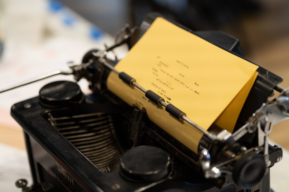 A vintage black typewriter with yellow paper loaded, showing a typed poem with partially legible text including the words 'alone,' 'body,' and 'promise of death.' The word 'Silent' is visible on the typewriter's body. 