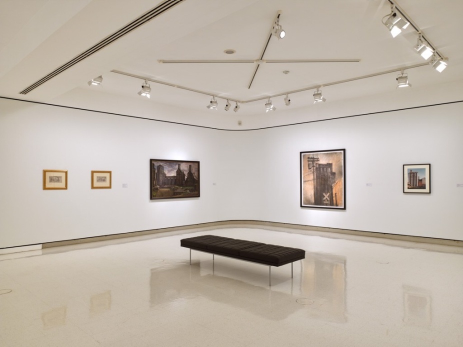 Installation view of Water, Grain, Steel: Industry and the Erie Canal at UB Anderson Gallery. The gallery features white walls and polished floors with five framed works depicting industrial scenes, including grain elevators and factories. A black bench sits in the center of the space beneath track lighting. 
