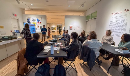 A group of participants seated at folding tables listen to two presenters during an Echoes workshop held inside the UB CFA Gallery, with the Martha Jackson in Paris exhibition visible on the surrounding walls.