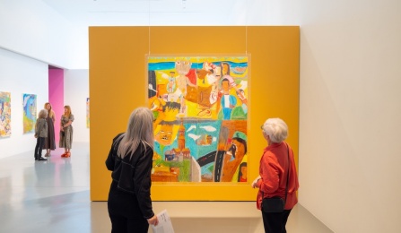 Two visitors view a large, colorful painting mounted on a mustard-yellow wall during the opening of bits by bits at UB Art Galleries. Additional visitors and artworks are visible in the background.