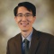 Headshot of a smiling man wearing grey, rectangular glasses, a light blue shirt, green tie, and brown suit jacket. 