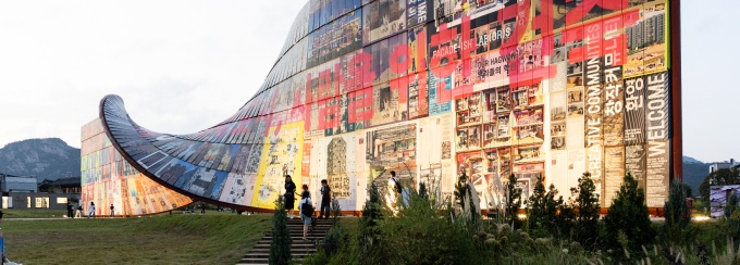 A large, curved installation with colorful images and text, set against a grassy landscape. 