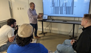 A person presents a "Vertical Fin Signage Facade Concept" on a screen to three attentive individuals. Transcribed Text: 수직 패널 사이니지 파사드 컨셉 [VERTICAL FIN SIGNAGE FACADE CONCEPT]. 