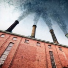 Smoke billows from three smoke stacks atop an aging, red brick industrial building. Stock photo via Canva. 