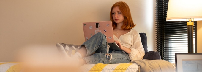 Student working on laptop on her bed in dorm room. 