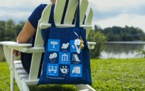Zoom image: Blue Campus Living Icon tote bag propped up on the back of an Adirondack chair with person sitting in it.