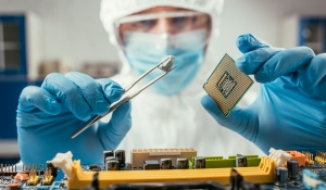 A worker in full lab gear (gloves, gown, facemask, hairnet) uses large forceps to place a small microchip into a computer motherboard. 