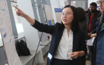 A student in formal gear points to her research poster while other students look on.