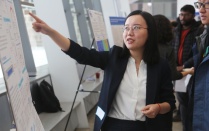 A student in formal gear points to her research poster while other students look on. 