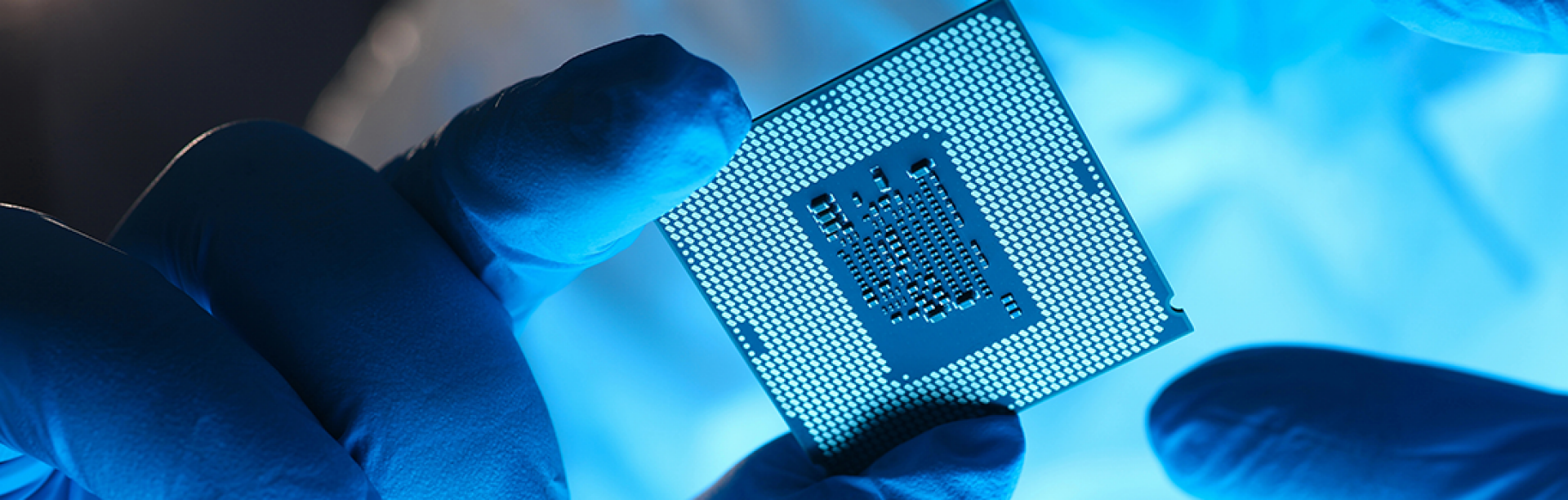 Gloved hands hold a square microprocessor chip, shown in close-up with its contact grid visible, illuminated by cool blue light. 
