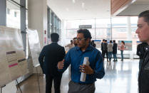 A crowd of students stand in the Davis Hall atrium. One student stands pointing to a poster and speaking while another examines the poster and listens to him. 