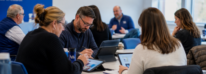 Faculty members collaborate in a university workshop, using tablets and notebooks while discussing ideas in small groups. 
