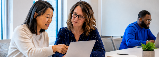 Two colleagues collaborate on a laptop in a bright office while a third works nearby; blank notebooks, papers and a UB mug sit on the table. 