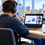 A person wearing headphones attends a virtual webinar on a laptop in a bright SUNY campus setting, with blue furnishings, study materials and a SUNY sign visible in the background. 