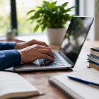 Person using a laptop at a desk with notebooks, books and a pen in a bright workspace, representing online teaching, learning and course development. 