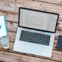 A laptop on a table. Next to the laptop on the left is a notepad with a pen and a glass of water. On the right of the laptop is a smartphone and a potted plant. 