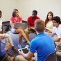 A group of students sitting in a circle and talking. On their laps are laptop computers. 