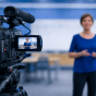 A video camera on a tripod records a person standing and speaking in a classroom, with the subject in focus on the camera screen and blurred in the background. 