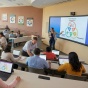 A classroom setting where a diverse group of students engages with digital learning materials that exemplify Universal Design for Learning (UDL) principles. Brightly lit with natural lighting, the room features accessible technology like adaptive devices and large screens displaying interactive content. 