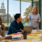 Diverse university staff collaborate around a conference table while discussing a change-management plan, with colorful notes arranged on a glass wall and a campus building resembling UB&rsquo;s Hayes Hall visible through the windows. 