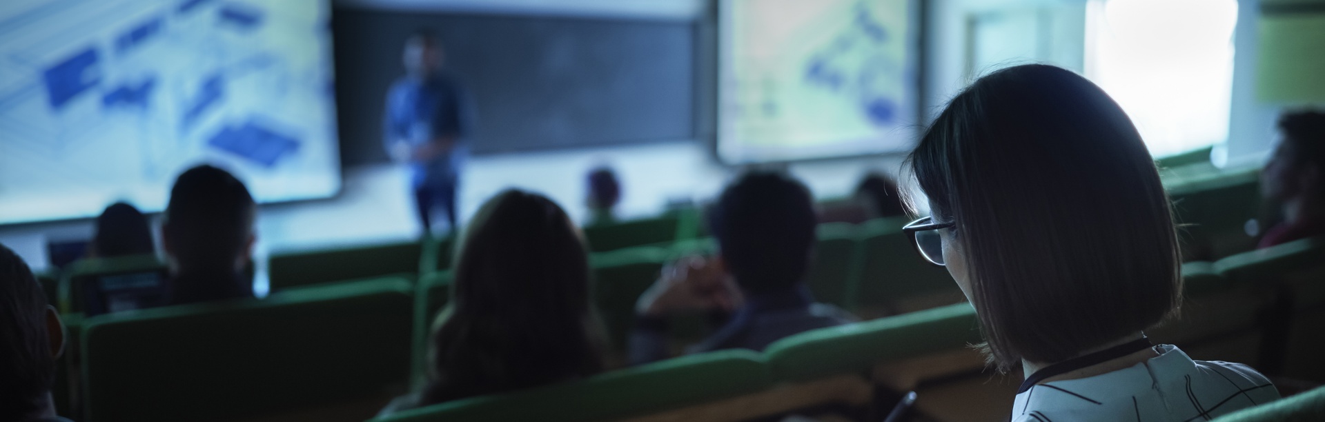 Students in a dark classroom with a faculty member presenting digital materials on a screen. 