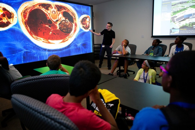 Zoom image: Large display showing image of internal organs with a room of teenagers and a presenter