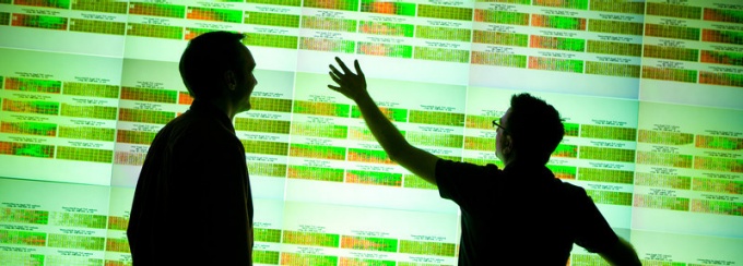 Two people are in shadow in front of a large tiled display wall with genomic information displayed on it. 