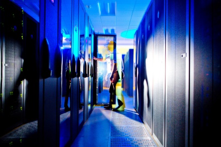 Zoom image: Male technician working on server in a rack with an open rack door in a row containing many racks of servers in a large data center