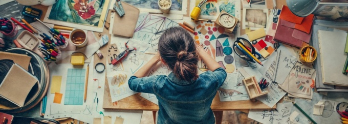 The Communities of Care sponsors art workshops in the community centered on the themes of the project. A top-down view of a young person with their hair in a bun and wearing a denim jacket, sitting at a messy desk covered in various art supplies and projects. The desk is cluttered with paintbrushes, colored pencils, sketches, paintings, and a small wooden model. The person is looking down at a piece of paper on the desk. 