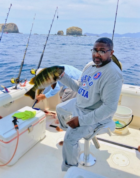 Kevin Smith, comfortably seated on a fishing boat, holds up a large fish in right hand. The fiberglass boat appears to be anchored on a sea of significance. In the background, large rock formations are nearby, with mountains in the distance. 