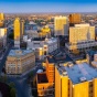 Birdseye aerial view of the City of Buffalo at sunset. The expansive urban scene faces south east. In the foreground, highlighted by the golden light of sunset, are several buildings in the downtown district, including the art deco style City Hall, and the ultra modern Court House. In the background, residential districts are visible in the distance. In the upper right corner, the Skyway winds out toward the shoreline of Lake Erie, showing the windmills of Lakawanna. The Boston hills appear in the distance. 