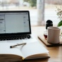 A laptop computer sits on a table. In front of the laptop is a book with a pen on it. To the right of the laptop is a flower pot, a cup of coffee, and a few other nicknacks. 
