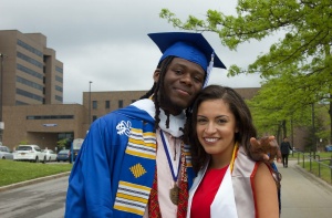 Graduates in cap and gowns standing in front of arena smiling on graduation day. 