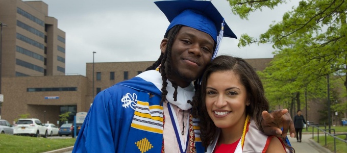 Graduates in cap and gowns standing in front of arena smiling on graduation day. 