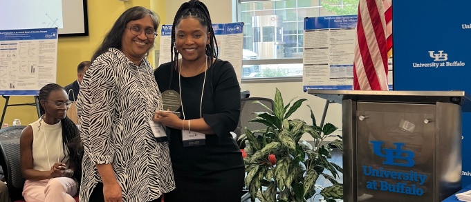 Director standing with faculty research mentor and holding a certificate in front of a UB logo background. 