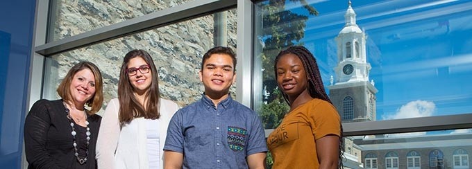 Four students standing in front of a large window overlooking a building with a white spire, with a blue sky in the background. 