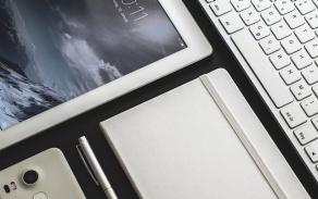 Photo of a white phone, notebook, keyboard, and tablet laid out on a black table. 