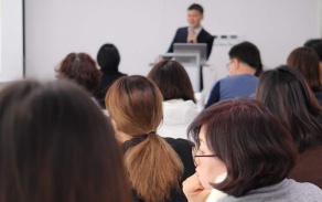 Photo of a seminar in which students (we see their heads from behind) watch a speaker at the front of a room. 