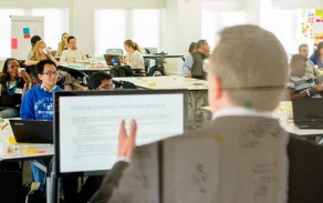 Photo of a presentation at UB's South Campus, taken of a presenter from behind, with attendees seated at tables and watching. 