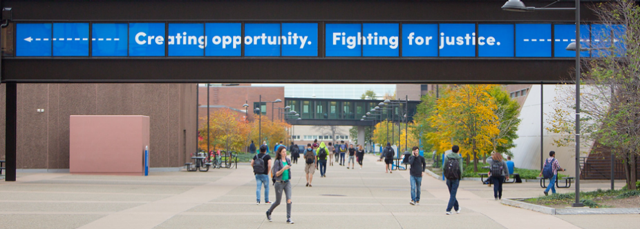 Students walk across UB&rsquo;s North Campus under a bridge banner reading, &ldquo;Creating opportunity. Fighting for justice.&rdquo;. 