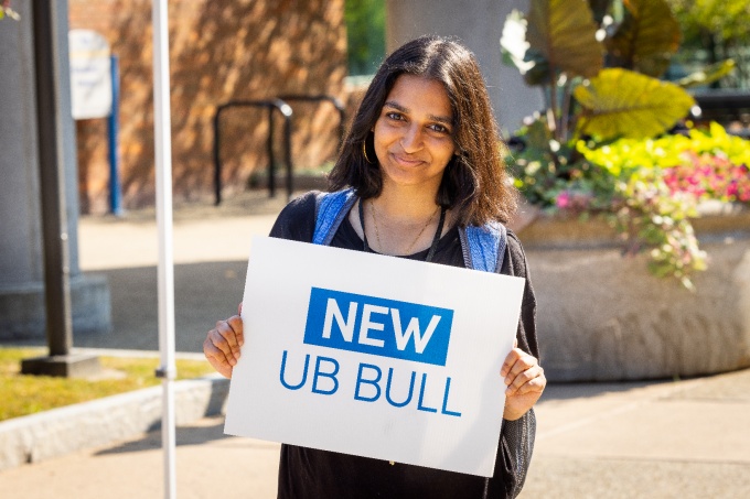 alt="New UB student holding a sign that says New UB Bull with North Campus in the background". 